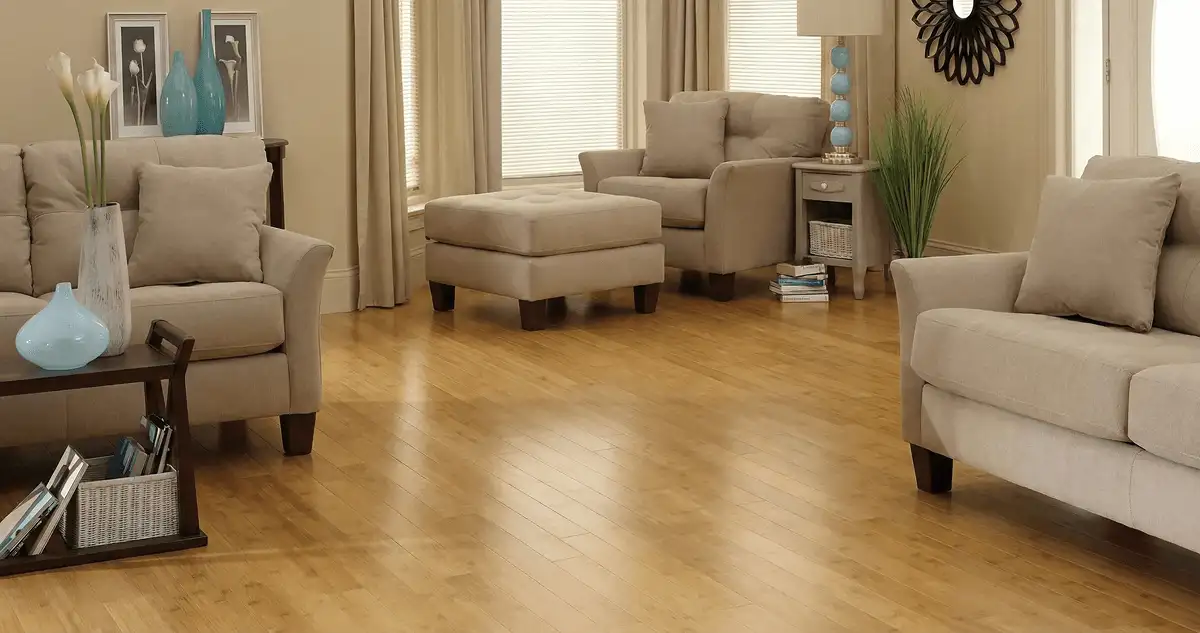 A living room with professionally installed bamboo flooring, beige sofas, a matching ottoman, a side table with a blue lamp, vases, framed pictures, and large windows with blinds and curtains in Boston, MA