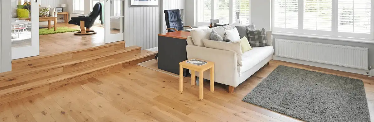 A modern living room with professionally installed laminate flooring, a beige sofa, a small side table, a gray rug, and large windows with white shutters in Boston, MA